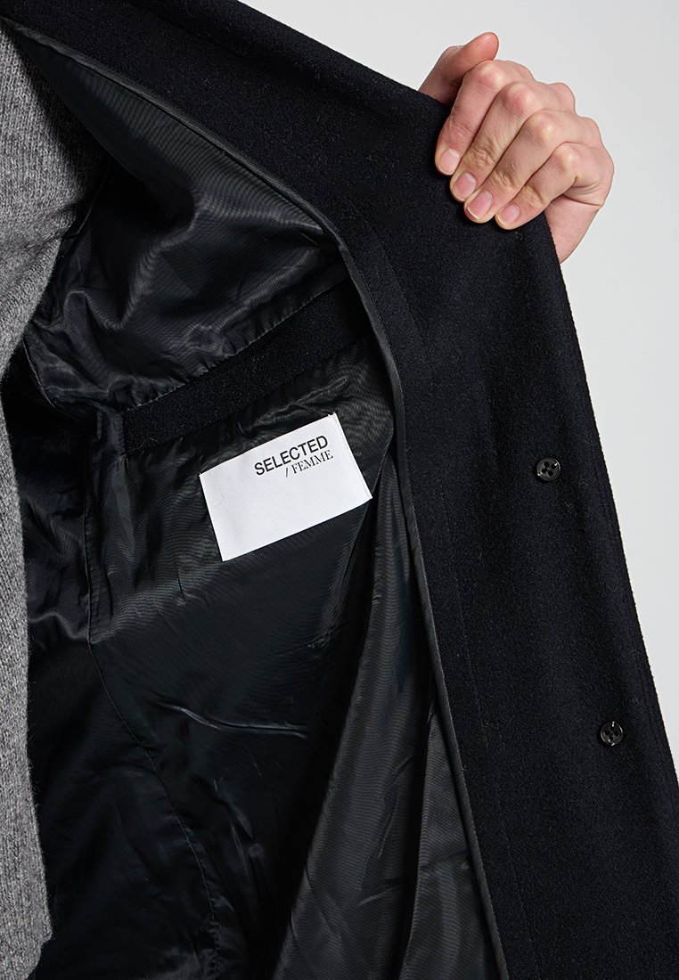 Intérieur de manteau noir, avec doublure noire lisse et une étiquette "SELECTED / FEMME". Bouton visible et tissu extérieur texturé.