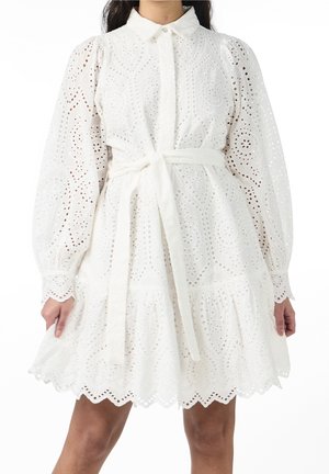 Vrouw draagt een witte eyelet kanten jurk met lange opgeblazen mouwen, een kraag, riem gestrikt in de taille en een geschulpte zoom.