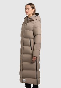 Long beige puffer coat with a hood, featuring a quilted design, black zippers, and a logo patch at the hem. Smooth, lightweight texture.