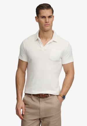 Mężczyzna w białej koszulce polo z krótkim rękawem, beżowych spodniach z plecionym brązowym paskiem i zegarkiem na rękę, stojący z jedną ręką w kieszeni.