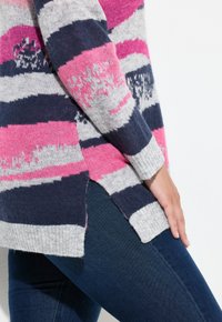 Persona che indossa un maglione a righe multicolore nei toni del rosa, blu navy e grigio, con uno spacco laterale, abbinato a jeans blu scuro.