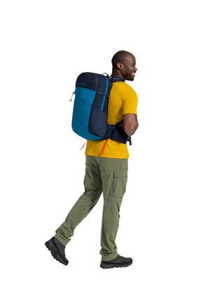 Mochila con una combinación de colores azul oscuro y verde azulado, con correas ajustables, un asa superior y acentos reflectantes. El usuario lleva pantalones cargo ligeros y botas.