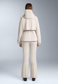 Skraćeni bež kaput s visokim ovratnikom i pojasom u struku, u kombinaciji s pripijenim bež hlačama koje imaju proširene nogavice i detalje sa patentnim zatvaračem na gležnjevima.