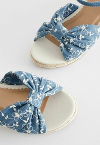 Sandales à bout ouvert avec des nœuds en tissu bleu brodés de fleurs blanches et des talons compensés beige sur fond uni.