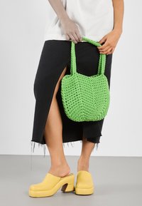 Green, woven bag with textured finish and two handles, paired with a black skirt featuring a side slit and yellow platform clogs.