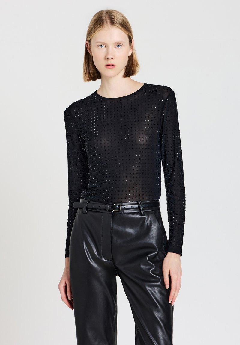 Schwarzes Langarm-Top aus durchsichtigem Material mit strukturiertem Punktdesign, kombiniert mit hoch taillierter schwarzer Lederhose und einem dünnen Gürtel.