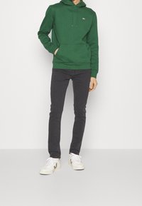 Sudadera verde con cordones ajustables, bolsillo frontal tipo canguro, combinada con jeans oscuros ajustados y zapatillas blancas con acentos beige.