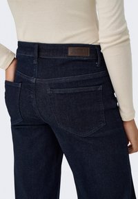 Vue de dos d'une personne portant un jean en denim bleu foncé taille haute et un haut à manches longues crème, main dans la poche arrière gauche.