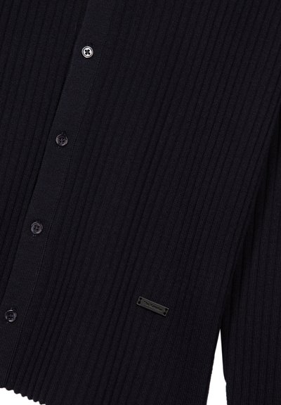 Cardigan noir en maille côtelée avec boutons sur le devant et une petite étiquette de marque rectangulaire près du bord inférieur du côté droit.