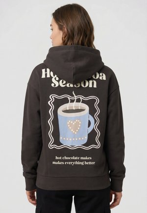 Vrouw draagt een donkere hoodie met stoom die opstijgt uit een blauwe mok en tekst "hot chocolate makes everything better" op de achterkant.