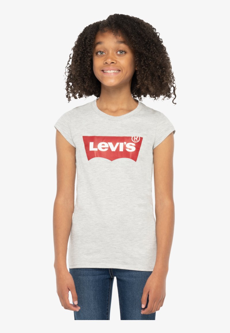 T-shirt gris clair à manches courtes avec un logo Levi's rouge prominent en design courbé, fabriqué en tissu doux, associé à un jean bleu.