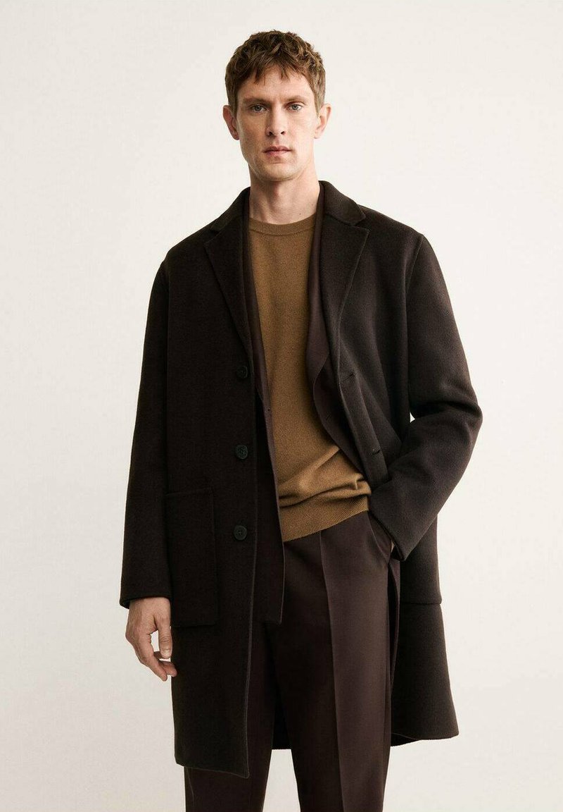 Cappotto in lana marrone con colletto a revers, dotato di tre bottoni, tasche laterali e vestibilità rilassata, indossato sopra un maglione marrone e pantaloni.
