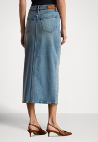 Ελαφρύ τζιν μίντι φούστα σε ανοιχτό μπλε χρώμα με ίσια σιλουέτα, δύο πίσω τσέπες και δερμάτινη ετικέτα στη μέση. Φορεμένη με καφέ τακούνια.