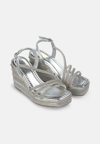 Sandalias de cuña plateadas con tiras entrelazadas, acabado metálico, plataforma texturizada y correa ajustable en el tobillo. Presenta acentos brillantes.