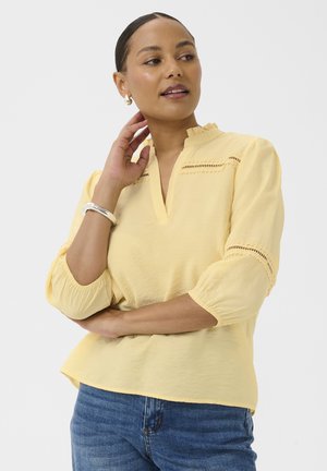 Kobieta z zaczesanymi do tyłu włosami, ubrana w jasnożółtą bluzkę z koronkowymi detalami i niebieskie dżinsy, pozująca z jedną ręką przy szyi.