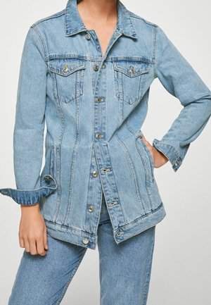 Person iført lyseblå denimjakke med knaplukninger, brystlommer og matchende jeans, stående med den ene hånd på hoften.