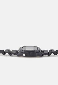 Digital watch with a square black case, rubber wavy strap, textured surface, and side buttons. LCD screen visible on top.