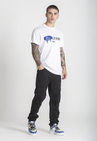 Mladý muž s tetovanými rukami stojí oblečený v bielom tričku s grafikou, čiernych cargo nohaviciach a modro-bielych teniskách s vysokým strihom.