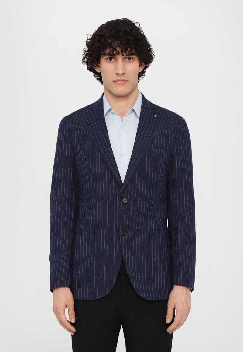Blazer a righe navy con vestibilità sartoriale, design monopetto, due tasche frontali e bottoni marroni. Abbinato a una camicia azzurro chiaro.