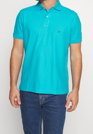 Turquoise polo shirt with a collar, two buttons, and short sleeves, paired with blue denim jeans. Features subtle logo detail on the chest.