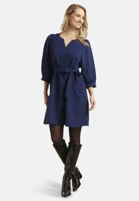 Marine jurk met een V-hals en pofmouwen, strakke taille met een stoffen riem; gecombineerd met zwarte kniehoge laarzen met hak. Soepele textuur.