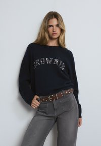 Pull marin avec "BROWNIE" en sequins, associé à un jean gris et une ceinture marron avec des accents étoilés. Tissu en mélange de coton et coupe décontractée.