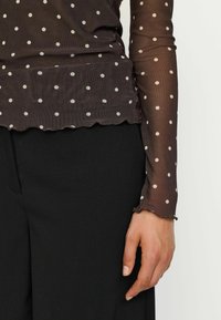 Genomskinlig brun långärmad topp med vita prickar, med en scalloped nederkant och manschettdetaljer, ihopparad med svarta byxor.