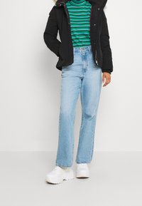 Person trägt hellblaue gerade Jeans, weiße klobige Sneaker, ein grün-blau gestreiftes Hemd und eine schwarze Daunenjacke mit Fellkragen.