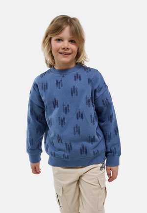 Jeune enfant aux cheveux blonds mi-longs porte un sweat bleu avec des motifs sombres de blé et un short cargo beige avec un petit motif noir.