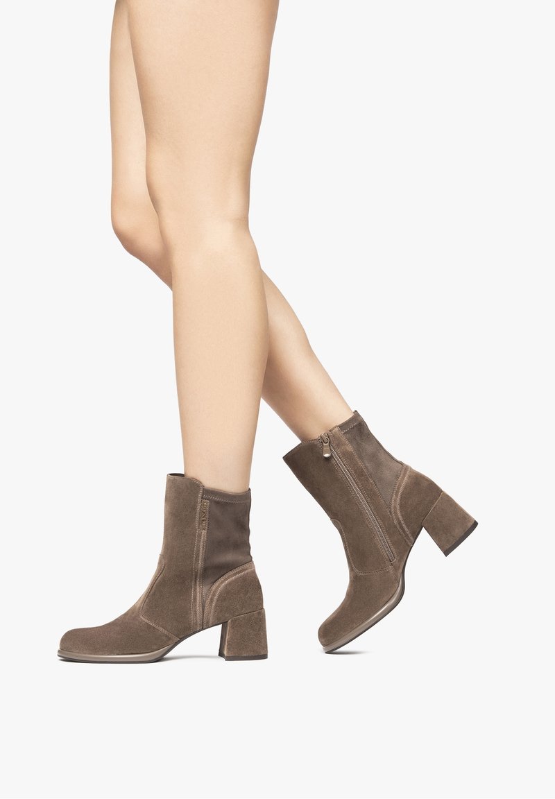 Jambes portant des bottes en daim marron mi-mollet avec talons carrés et fermetures éclair latérales, montrées en position de marche sur un fond uni.