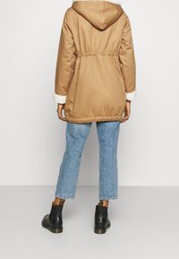 Osoba stojící s otočením zády, oblečená v béžovém dvoudílném kabátu s bílými fleecovými manžetami, zkrácených modrých džínsech a černých kotníkových botách.