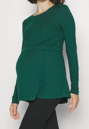 Personne enceinte portant un haut de maternité vert foncé à manches longues associé à un pantalon noir, posée devant un fond uni.
