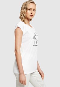 Hvid bomulds t-shirt med korte rullede ærmer, der har en minimalistisk sort linjetegning af et ansigt og teksten "Undervurder aldrig en..."