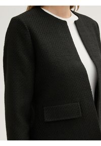 Chaqueta negra texturizada con un cuello redondo y sin botones. Presenta un bolsillo tipo parche en la parte frontal y una blusa blanca de canalé debajo.