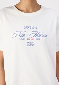 Balta medvilninė marškinėliai su apvalia iškirpte. Priekyje yra mėlynai siuvinėti tekstai: "GANT 1949", "New Haven" ir "Sailing Crew", taip pat dekoratyvinės vėliavos.