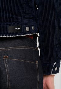 Granatowa kurtka z korduroya z miękką białą podszewką; posiada srebrne guziki oraz czarną metkę z napisem "Pepe Jeans London" w dolnej lewej części.