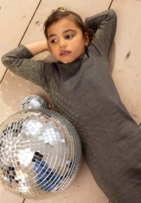 Glänzendes, dunkelgraues Kleid mit metallischen Fäden, figurbetont. Neben einer kugelförmigen Discokugel mit Spiegelfliesen, auf einem Holzfußboden.