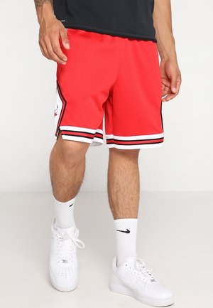 Homme portant un short de basket rouge avec des rayures noires et blanches, des chaussettes blanches Nike et des baskets blanches Nike, debout sur un fond uni.