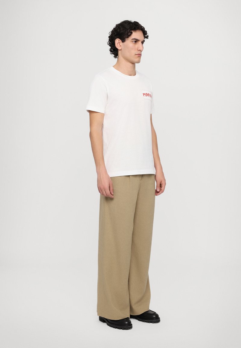 White cotton t-shirt with red text, paired with beige wide-leg trousers and black chunky boots. Man stands sideways against a plain background.