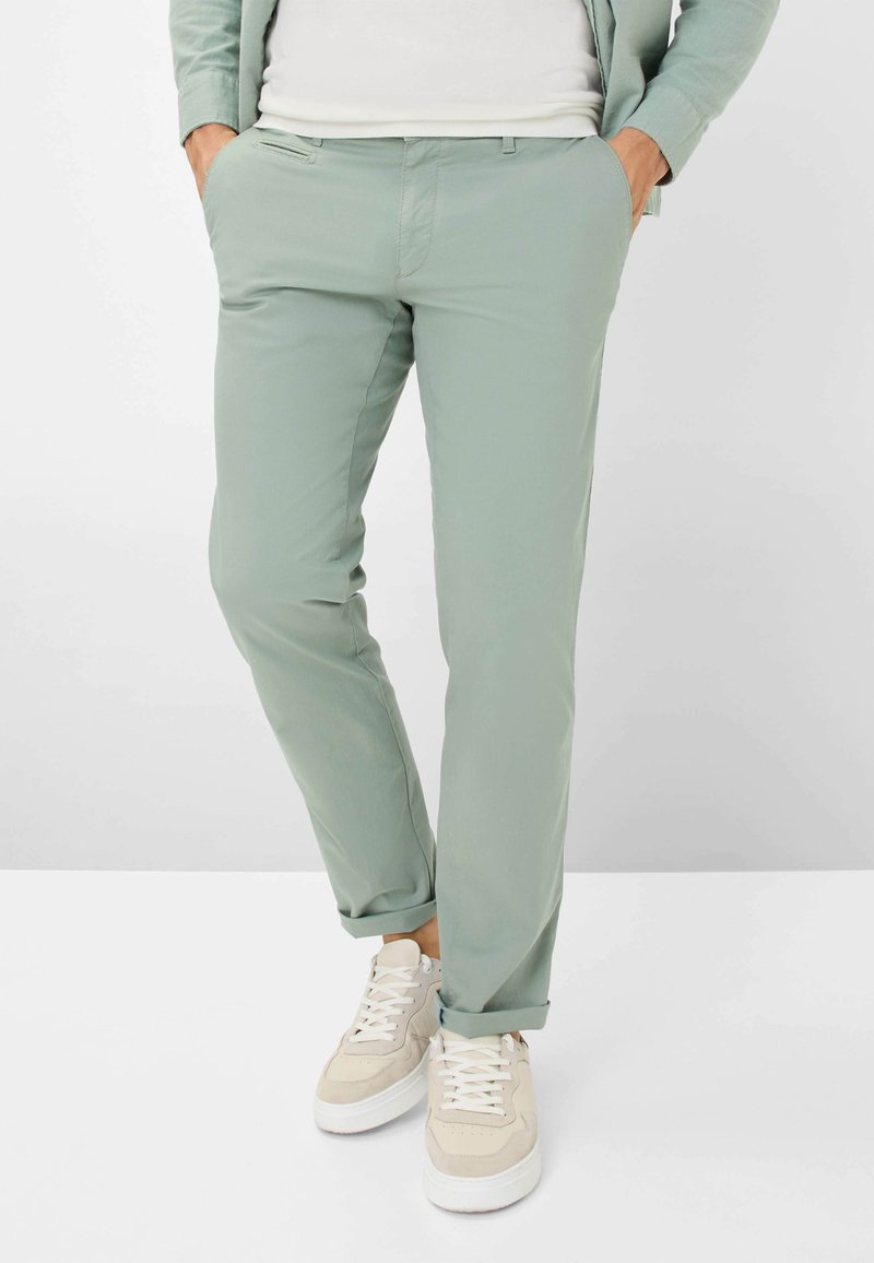 Helle grüne Hose aus glattem Stoff mit schmaler Passform, vorderen Taschen und aufgeschlagenen Säumen, getragen mit beigen Sneakers.