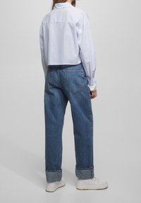 Donna che indossa jeans blu a pantalone corto, sneakers bianche e una maglietta corta a righe azzurre, in piedi di spalle contro uno sfondo semplice.