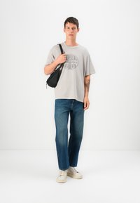 Grå oversized t-skjorte med et sirkulært logo, blå straight-leg jeans og kremfargede sneakers; holder en liten svart crossover-veske.