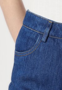 Jeans de ganga azul escuro com um bolso frontal e passantes para cinto, com costura dourada em contraste ao longo das costuras e bordas dos bolsos.