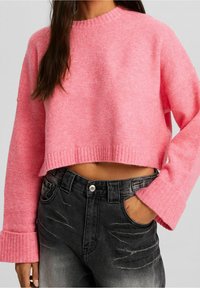 Femme portant un pull court rose avec de larges poignets et un jean taille haute noir délavé, main dans la poche, sur un fond uni.