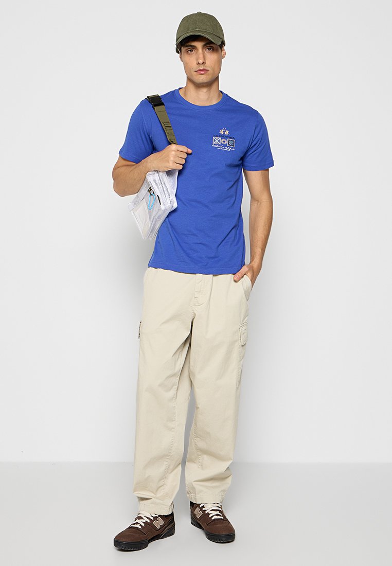 Blaues Baumwoll-T-Shirt mit einem Logo, beige Cargo-Hose und braunen Schuhen. Leichte Umhängetasche ist durchsichtig mit bunten Reißverschlüssen. Grüner Hut.