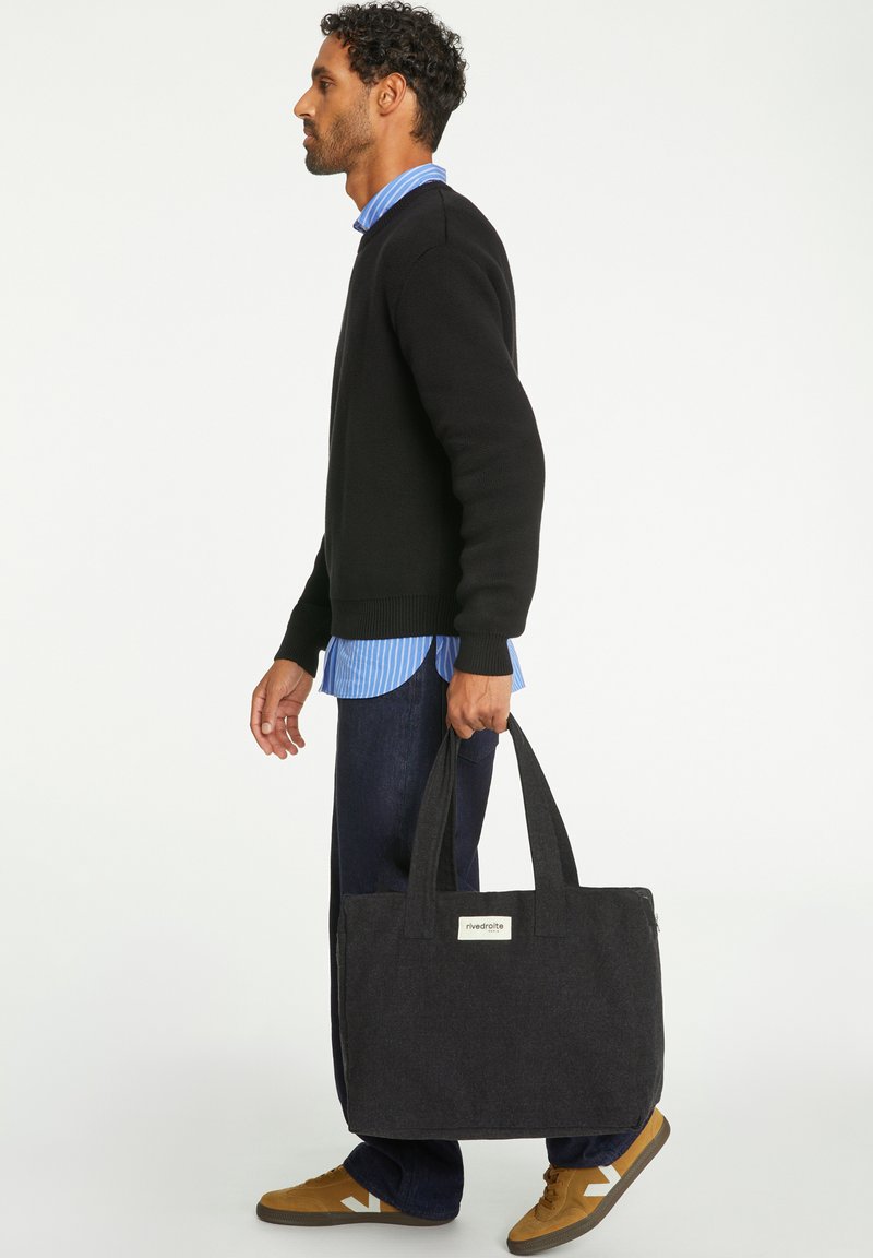 Sac fourre-tout noir en tissu avec deux poignées solides, présentant une étiquette à l'avant. Personne portant un pull noir, une chemise à rayures bleues et un jean en denim foncé.