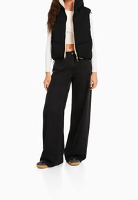 Black puffer vest with hood, zippered front, and beige lining, paired with high-waisted, wide-leg black jeans and black shoes with brown soles.