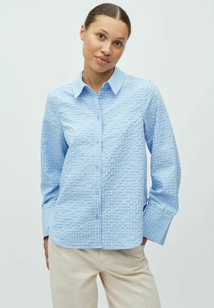 Ženska oblečena v svetlo modro teksturirano srajco z gumbi in ovratnikom ter bež hlače, stoji ob preprostem svetlem ozadju.