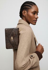 Smeđa torbica sa uzorkom zmijske kože i zlatnom lančićem, koja ima kopču i pravokutni oblik, nošena preko svijetlosmeđeg kaputa.