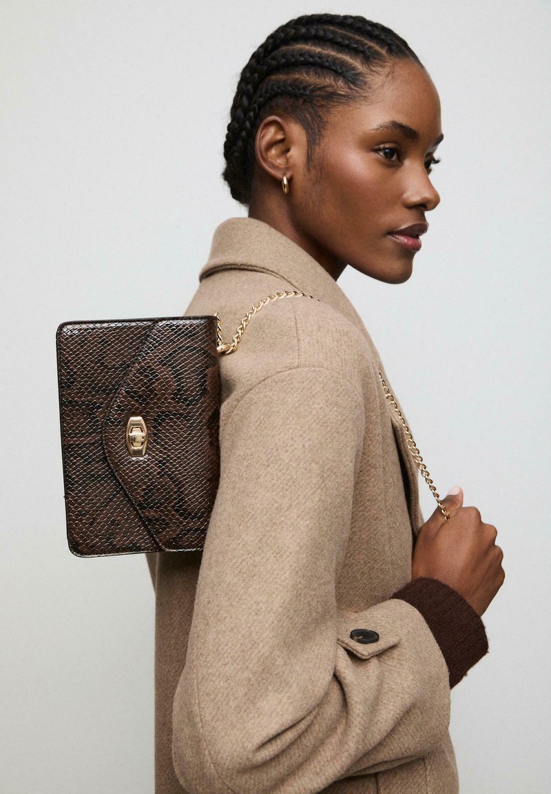 Smeđa torbica sa uzorkom zmijske kože i zlatnom lančićem, koja ima kopču i pravokutni oblik, nošena preko svijetlosmeđeg kaputa.